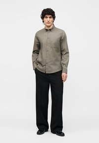 Homme aux cheveux bouclés portant une chemise olive boutonnée, pantalon noir à jambes larges et chaussures noires, debout avec une main dans la poche.
