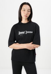 T-shirt en coton noir avec une coupe ample, arborant un texte stylisé blanc "Doon Jeans" et un design graphique subtil, porté avec un pantalon noir.