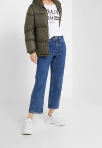 Olivově zelená péřová bunda s kapucí, bílý triko s grafikou, modré džíny s vysokým pasem a rovnými nohavicemi a bílé tenisky.