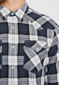 Chemise à manches longues en flanelle à carreaux bleu marine et blanc, avec poches poitrine, boutonnage frontal et motifs à carreaux contrastants. Texture en tissu de coton.