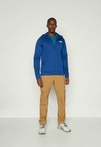 Μπλε φούτερ με φερμουάρ και μακριά μανίκια, με ψηλό γιακά, συνδυασμένο με καφετί cargo παντελόνι και αθλητικά παπούτσια ανοιχτού χρώματος. Ελάχιστο branding ορατό.