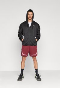 Sweat à capuche noir zippé avec capuche et accents blancs, associé à un short bordeaux avec des contours roses. Chaussures de sport noires avec détails verts.