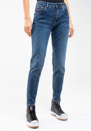 Vaqueros ajustados de denim azul con un sutil desgaste, cinco bolsillos y trabillas para cinturón. Combinados con zapatillas altas negras con suelas blancas.