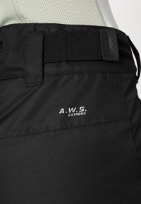 Musta tekniset housut, joissa on leveä elastinen vyötärö, joka on tekstuuroitu, ja valkoinen logo, jossa lukee "A.W.S. EXTREME." Läheltä nähtynä.
