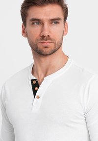 Muž s krátkými hnědými vlasy a vousy, oblečený v bílém dlouhém rukávu Henley tričku se třemi dřevěnými knoflíky a černým vnitřním límcem.