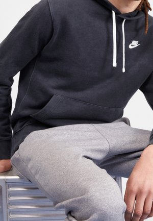 Person trägt einen schwarzen Nike-Hoodie mit weißen Kordeln und graue Jogginghose, sitzt auf einer metallischen Bank oder einem Hocker.