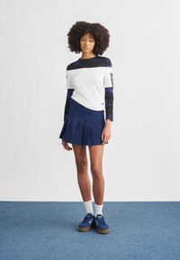 Hvit og blå lagvis langermet topp med svarte detaljer på armene, kombinert med en marineblå plissert skjørt og blå joggesko, satt mot en hvit bakgrunn.