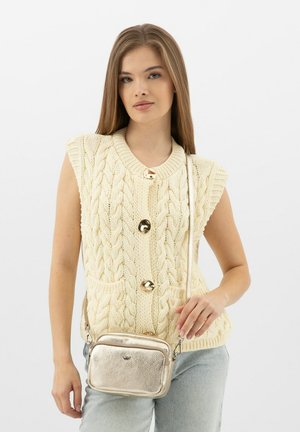Gilet en tricot torsadé crème avec des boutons dorés, manches courtes et deux poches avant. Le modèle porte un petit sac bandoulière en métal argenté.