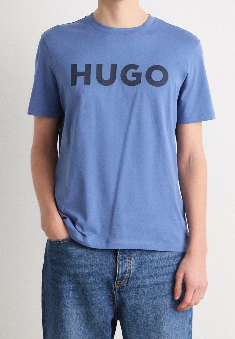 Personne portant un T-shirt bleu clair avec "HUGO" imprimé en grandes lettres noires, associé à un jean bleu, debout devant un fond uni.