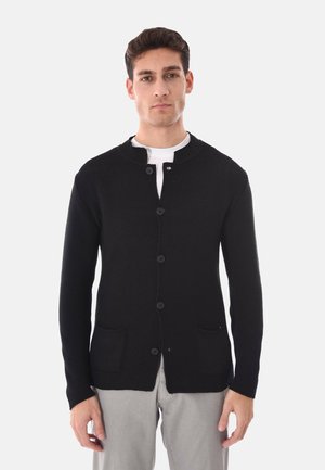 Uomo che indossa un cardigan nero con bottoni e due tasche frontali sopra una camicia bianca e pantaloni grigio chiaro, in piedi contro uno sfondo semplice.