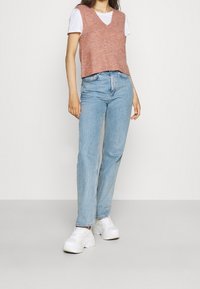 Person in einem weißen T-Shirt unter einem rosaroten ärmellosen Pullover, hellblauen geraden Jeans und weißen plateausneakern, die an einer schlichten Wand stehen.