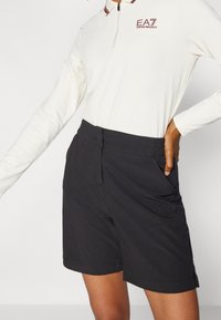 Osoba oblečená v černých na míru šitých šortkách a bílém polo tričku s dlouhým rukávem, které má čtvrtinový zip a červené logo na hrudi.