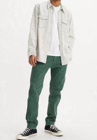 Chemise boutonnière gris clair avec deux poches poitrine, associée à un pantalon vert et des baskets noires avec des bouts et des lacets blancs.