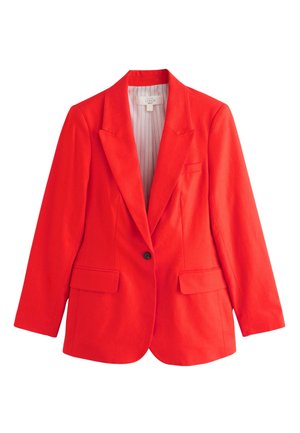 Kvinde med langt mørkt hår iført en rød dragt med en enkelt-knaps blazer og bukser med vide ben, stående indendørs mod en ensfarvet væg.