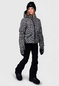 Νέος άνθρωπος που φοράει μπουφάν με λεοπάρ σχέδιο, μαύρα παντελόνια χιονιού, ταιριαστά γάντια, μαύρες μπότες και μαύρο σκουφάκι, στέκεται σε χαλαρή στάση.