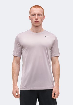 Jeune homme aux cheveux roux courts portant un t-shirt de sport Nike gris clair à manches courtes et un short noir, debout devant un fond uni.
