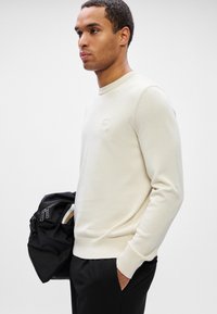 Ein Mann in einem cremefarbenen Pullover und schwarzen Hosen hält in einer Hand eine schwarze Tasche, während die andere Hand in der Tasche steckt, und steht vor einem schlichten grauen Hintergrund.