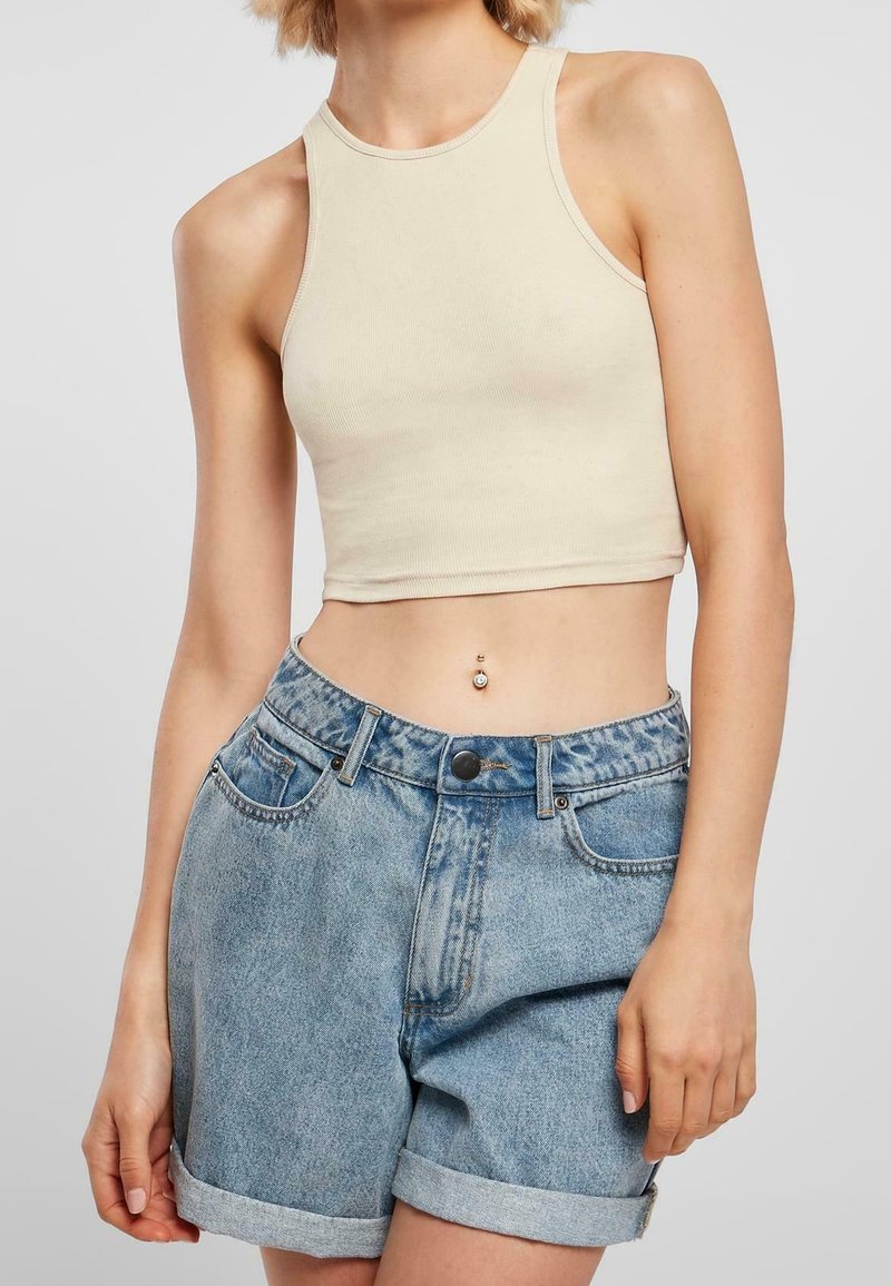 Personne portant un crop top beige sans manches et un short en jean taille haute retroussé, montrant un piercing au nombril.