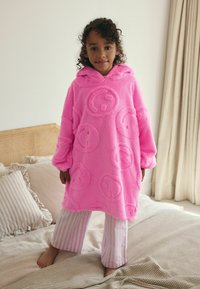 Poncho en polaire rose avec des motifs de visages souriants, capuche et manches surdimensionnées. Associé à un pantalon de pyjama à rayures. Tissu doux et texturé.
