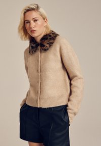 Cardigan beige en tricot avec un col en fausse fourrure texturé à motif léopard, doté d'une fermeture à boutons sur le devant et d'un ourlet côtelé. Porté par-dessus un short en cuir noir.