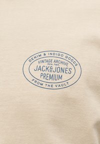 Μπεζ ύφασμα με μπλε οβάλ λογότυπο που γράφει «Denim & Indigo Goods Vintage Archive Jack & Jones Premium From the Vault».