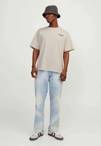 Homme portant un t-shirt beige "COSMOS", un jean bleu clair usé, un chapeau de seau et des baskets, se tenant devant un fond blanc uni.