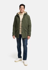 Grüne Cordjacke mit einer fellgefütterten Kapuze, Druckknöpfen auf der Vorderseite und Taschen; getragen über einem beigen Pullover und kombiniert mit blauen Jeans und weißen Schuhen.