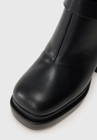 Fekete bőrcsizma négyszögletű orral, látható varrással és vastag talppal, sima világos háttér előtt.