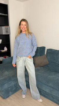 Pulover tricotat albastru deschis cu pete, asortat cu blugi largi din denim deschis. Fundalul include o canapea verde și o unitate de rafturi.