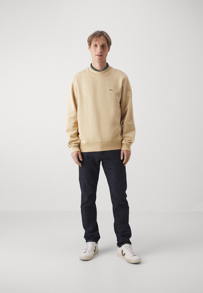Beige Sweatshirt mit gerippten Bündchen, Rundhalsausschnitt und kleinem Logo. Kombiniert mit dunkelblauen Jeans und weißen Sneakers mit grünen Akzenten.