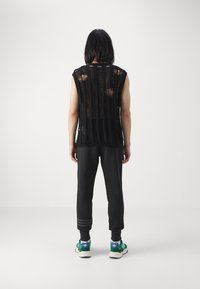 Top in rete nera senza maniche con fessure verticali, abbinato a pantaloni della tuta neri. Sneakers verdi con dettagli viola completano l'outfit.