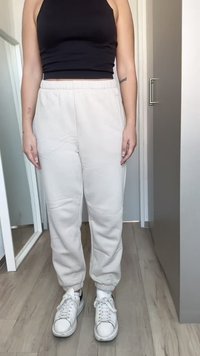 Beige sweatpants with elastic waistband and cuffs, featuring a smooth texture. Paired with white sneakers, standing indoors.