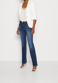 Blazer blanco con acentos dorados, camisola de encaje beige, jeans rectos azul oscuro y sandalias blancas de tacón abierto. Tejido suave y diseño ajustado.