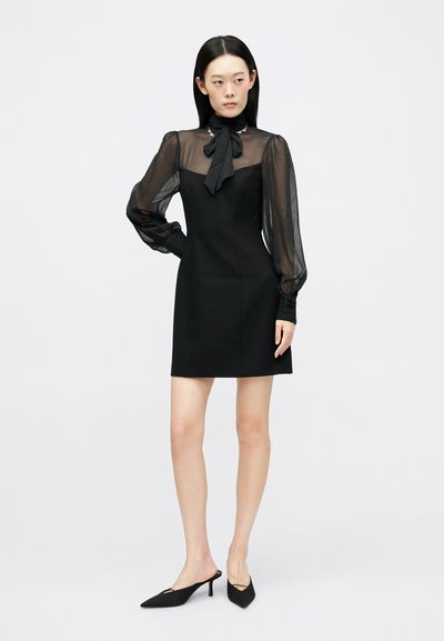 Μαύρο φόρεμα με διάφανα μακριά μανίκια και γιακά με φιόγκο. Στενή σιλουέτα, μαλακή υφή, έως το γόνατο, συνδυασμένο με μαύρες μυτερές μούρες.