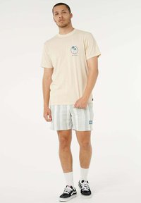 T-shirt beige chiaro con grafica di palma, abbinata a pantaloncini chiari a righe. Sneakers nere, calzini bianchi alla caviglia. Design minimalista e vestibilità casual.