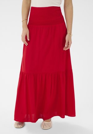 Žena má na sebe dlhú červenú sukňu so širokým nariaseným pásom, biely top, zlaté náramky a béžové pletené baleríny na bielom pozadí.