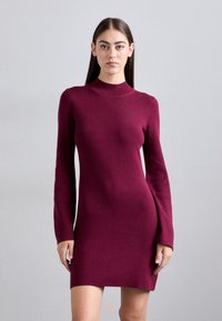 Fiatal nő egy egyenes, világosszürke háttér előtt áll, testhezálló, hosszú ujjú bordó kötött ruhát viselve.