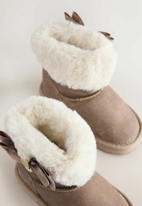 Bruine suede enkellaarzen met een pluizige witte nepleren voering, voorzien van twee donkerbruine lintaccenten aan de zijkant. Zachte, afgeronde vorm.