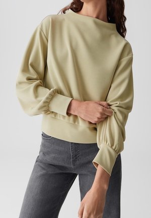 Persoon draagt een lichtbeige top met lange mouwen en pofmouwen en donkergrijze spijkerbroek, en past de rechtermouw aan met de linkerhand.