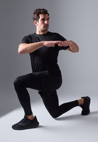 Črno športno majico in legice, model v pozi izpadnega koraka, obut v črne tekaške čevlje. Gladka tekstura, telesu prilegajoča zasnova.