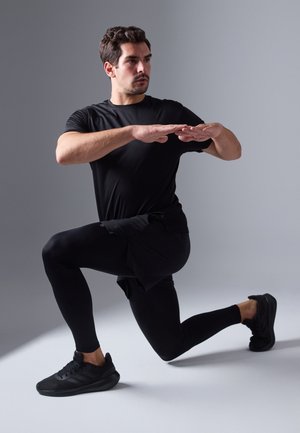 Μαύρη αθλητική μπλούζα και κολάν, μοντέλο σε θέση lunges, φορώντας μαύρα αθλητικά παπούτσια. Λείος υφή, εφαρμοστό σχέδιο.