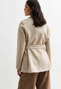 Abrigo de mezcla de lana beige con un diseño ajustado, cintura ceñida y textura suave. Presenta un respaldo entallado con costuras estructuradas.