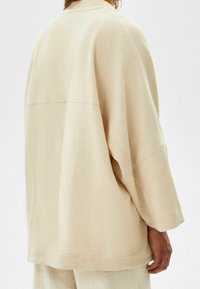 Beige oversized skjorta i strukturerad, ribbad tyg, med en lös passform, vida ärmar och hög hals, sedd från baksidan.