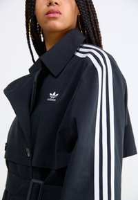 Manteau noir avec un grand col, une ceinture en tissu et un détail à trois bandes blanches Adidas sur les manches, rehaussé d'un petit logo.