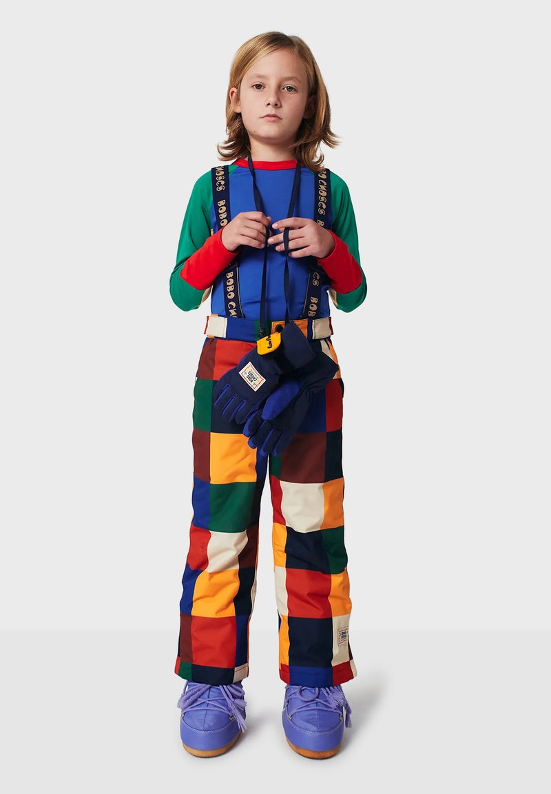 Farverigt skioutfit med patchwork-snebukser i rød, grøn, gul og brun, en blå top og blå handsker med orange detaljer.