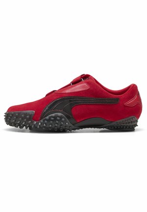 Chaussure de sport en daim rouge avec bande en cuir noir, semelle noire à crampons et design sans lacets sur fond blanc.