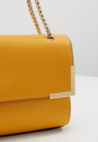 Žlutá kabelka s texturou a hladkým povrchem, s řetízkovým popruhem ze zlata a geometrickým kovovým detailem na přední straně.