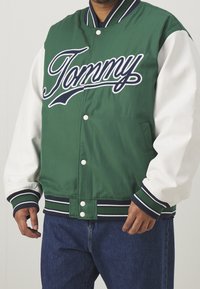 Zielono-biała kurtka varsity z haftem "Tommy" na piersi, wyposażona w zatrzaski z przodu, ściągacze na mankietach oraz paskowany pas.