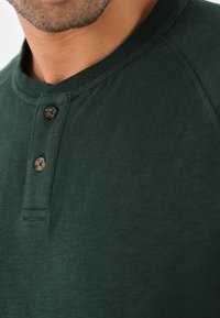 Tumedat rohelised henley-särk, millel on ümmargune kaelus, kahe nupuga detail ja pehme, tekstuuriga kangas. Omab raglan-varrukaid ja puhtat viimistlust.