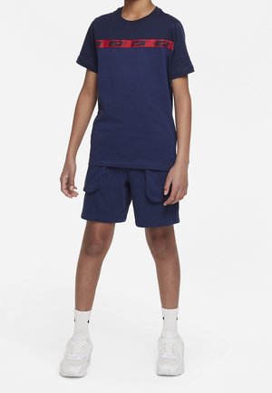 Chłopiec w granatowej koszulce Nike z czerwoną paską logo, granatowych spodenkach z przednimi kieszeniami, białych skarpetkach i białych butach sportowych stoi prosto.